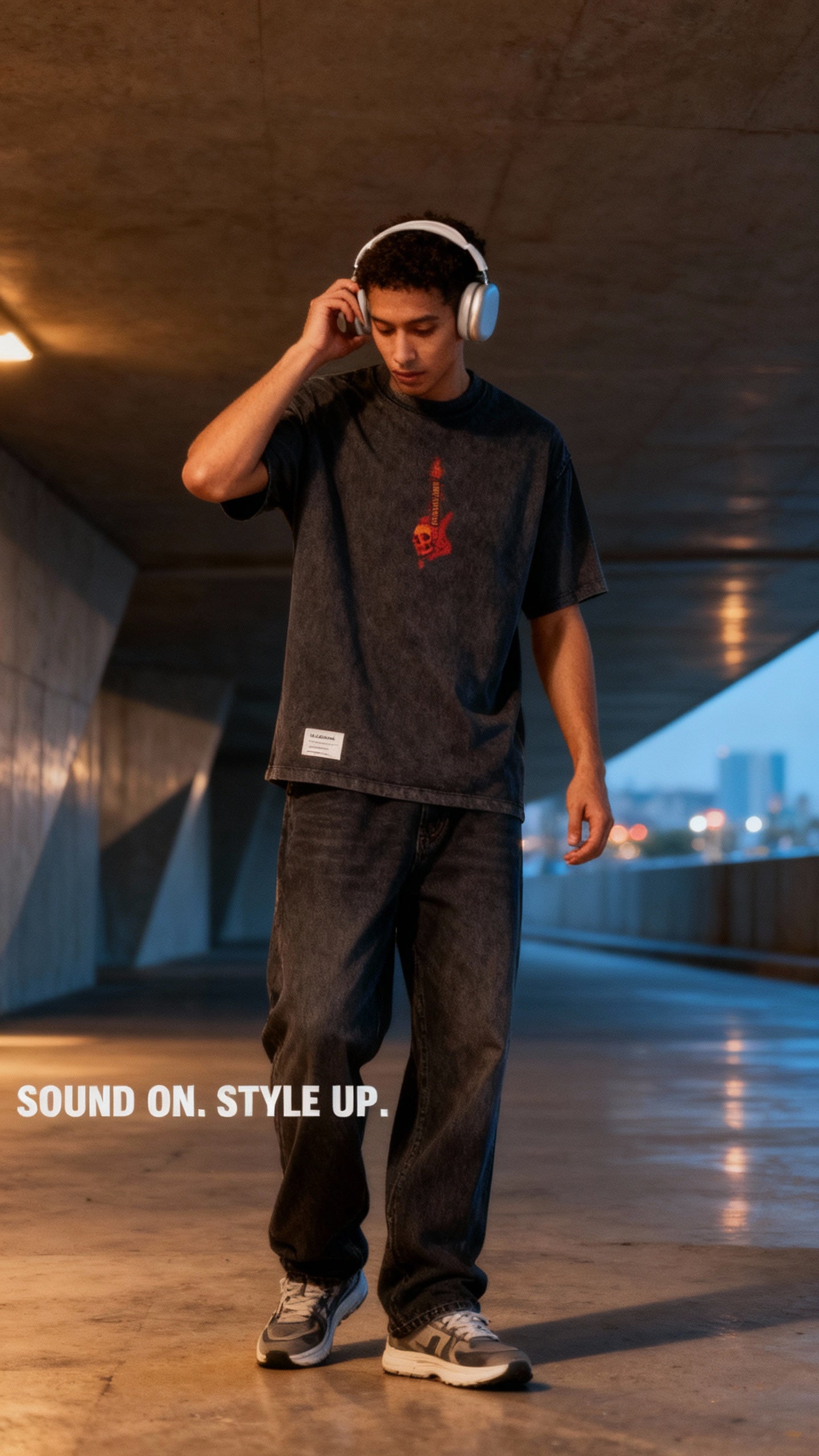 Giovane uomo con cuffie wireless bianche, t-shirt nera con chitarra rossa, jeans e sneakers grige in tunnel urbano al tramonto