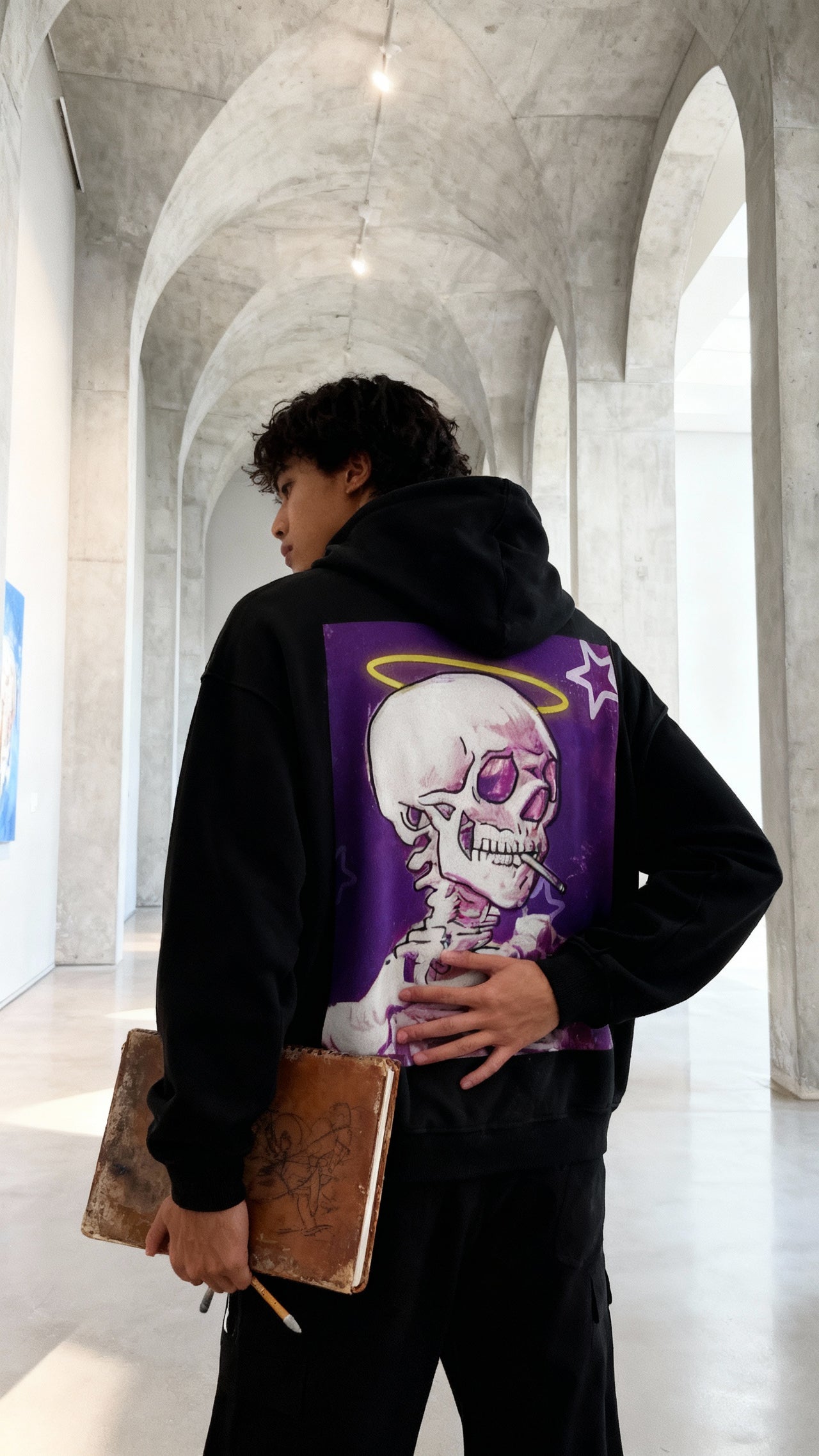 Giovane con felpa nera con stampa teschio e aureola gialla, in galleria d'arte moderna con archi in cemento